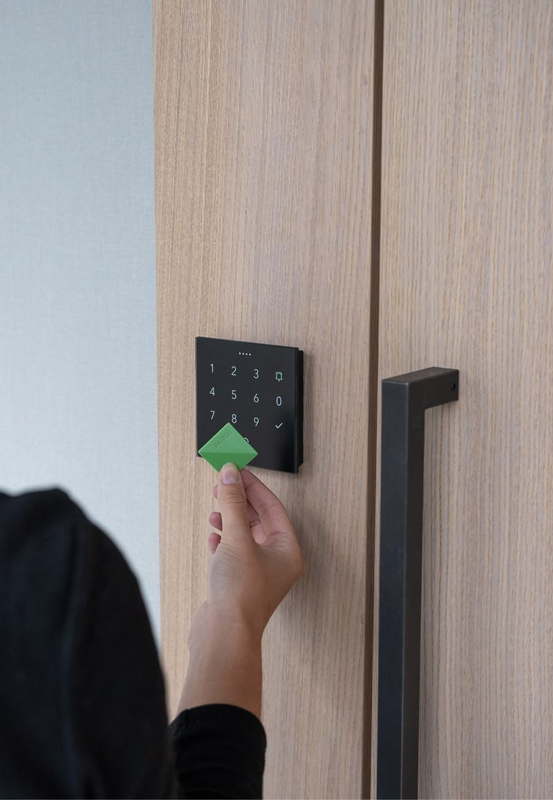 Dłoń przykłada zielony breloczek do czytnika Loxone NFC Code Touch przy drzwiach z jasnego drewna